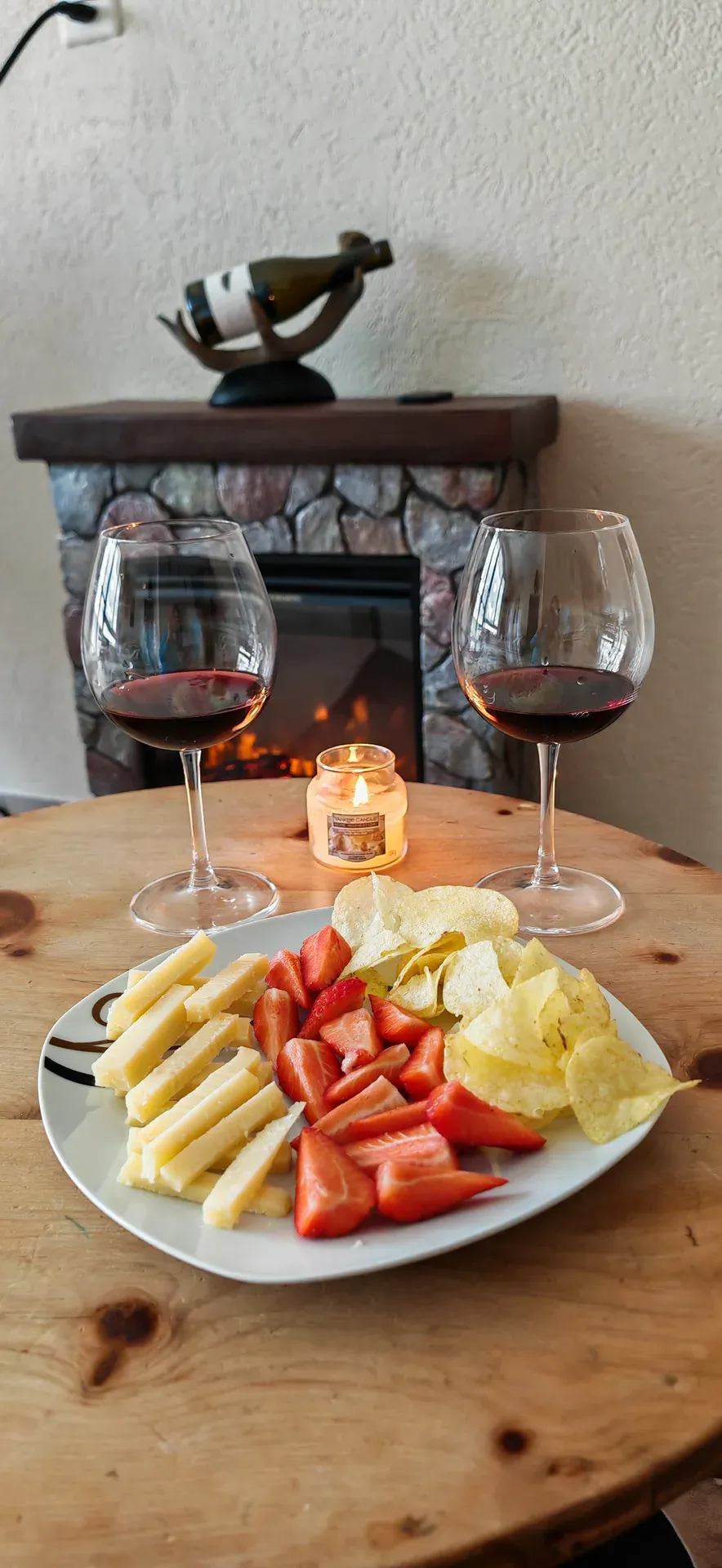 Zwei Gläser Rotwein, Käse, Erdbeeren und Chips, dahinter ein Kamin