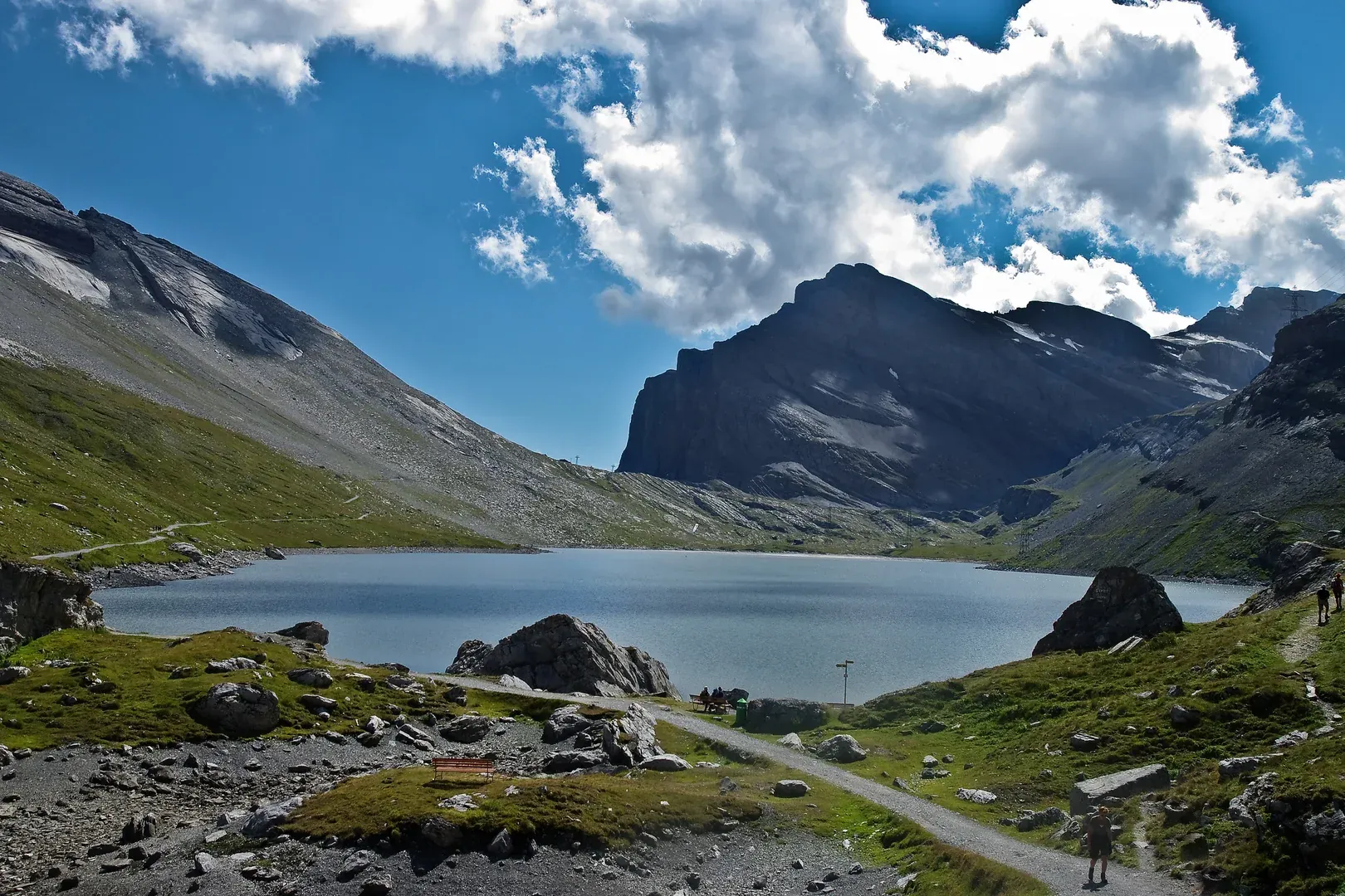Bergsee zwischen Geröllhängen, ein Kiesweg und ein paar Wanderer