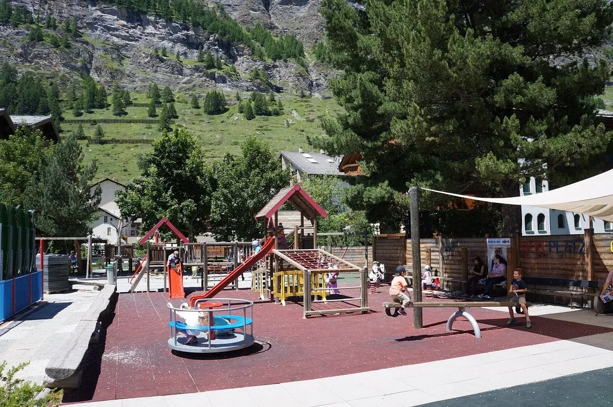 Spielplatz mit roter Rutsche und Karussell, Kinder und ein Berghang dahinter