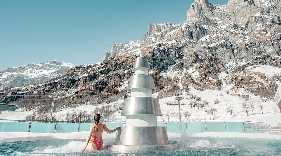 Person im Aussenbecken neben einem Metallbrunnen, dahinter schneebedeckte Felswände