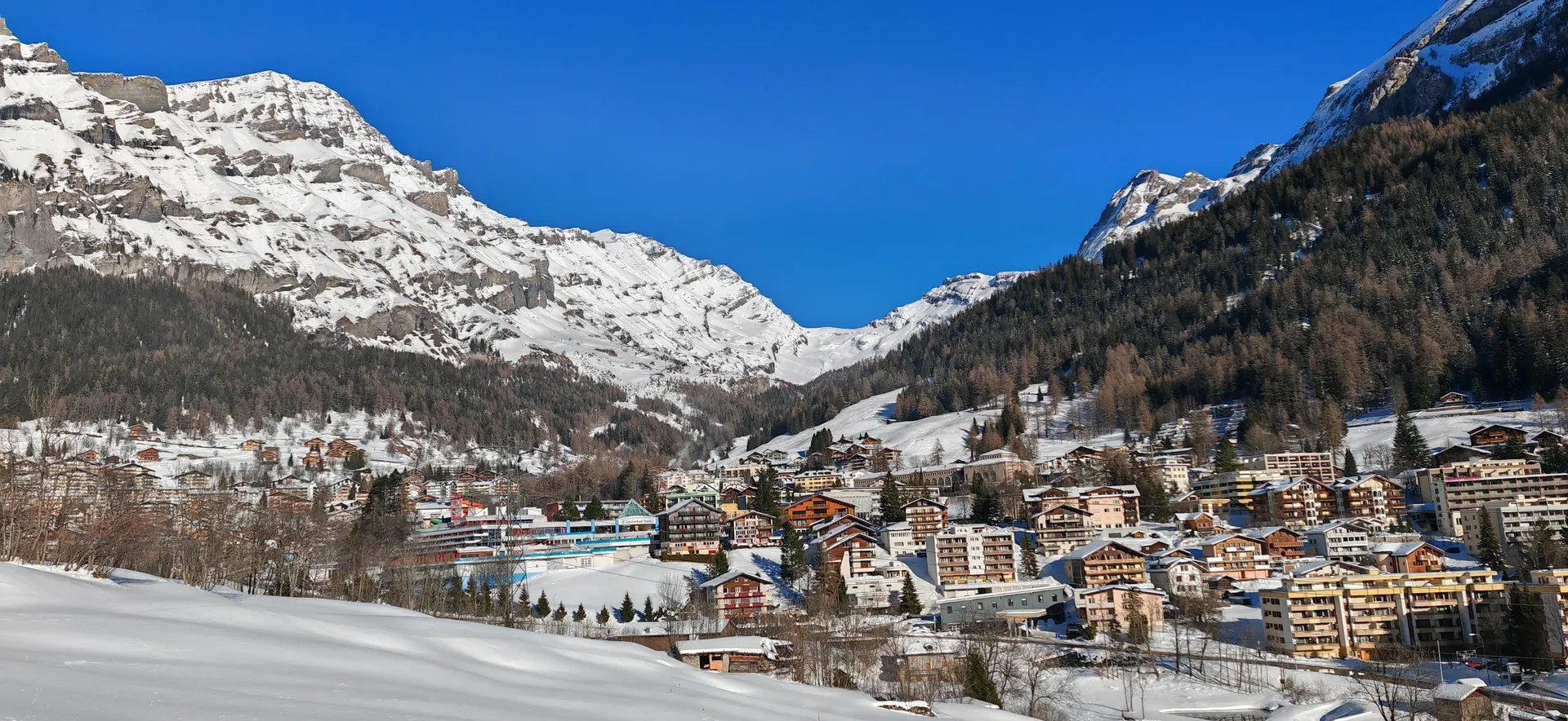 Verschneites Leukerbad im Tal, Holzhäuser und ein langes helles Gebäude vor Felswänden
