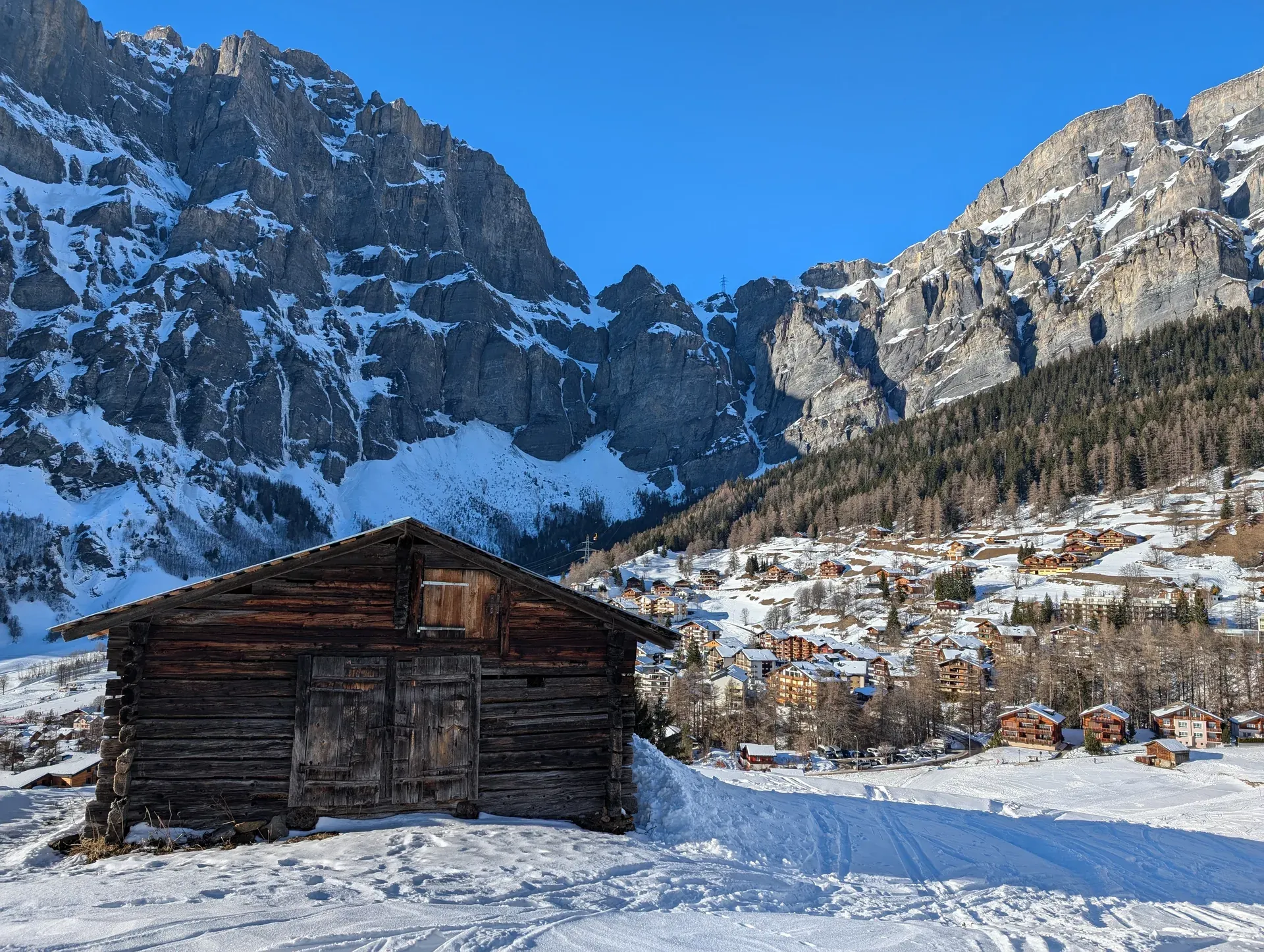 Dunkle Holzhütte im Schnee, dahinter ein Bergdorf und Felswände