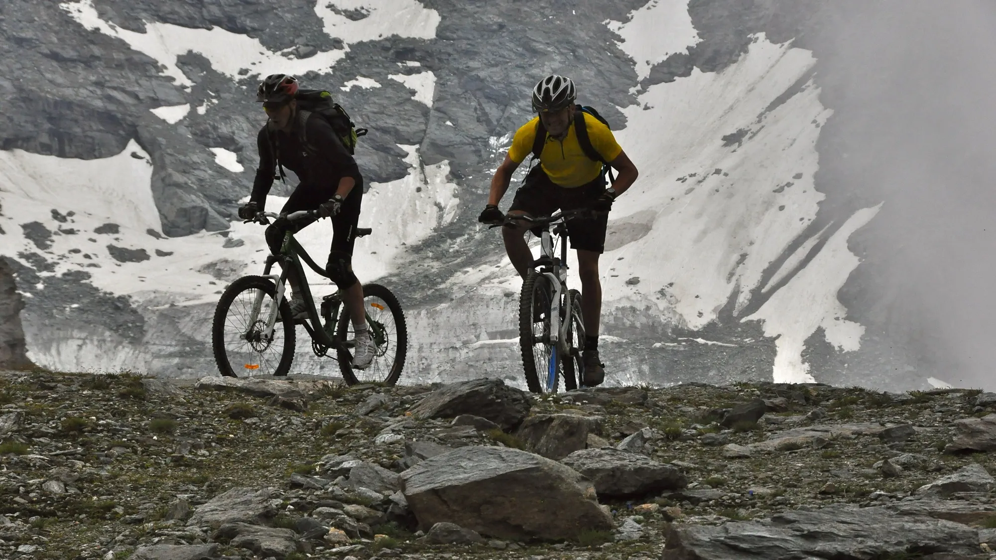 Zwei Mountainbiker fahren auf einem steinigen Weg vor einer Felswand mit Schneefeldern