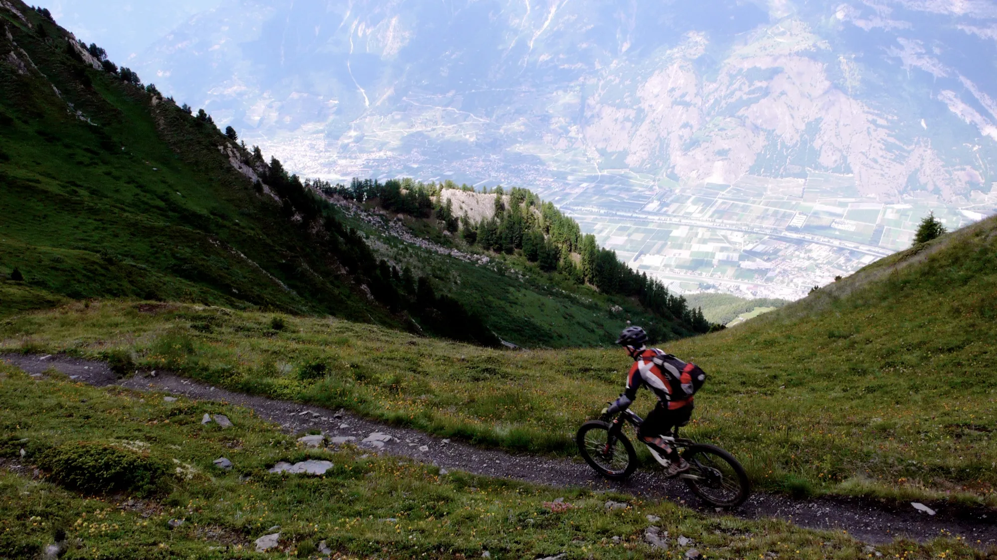 Mountainbiker auf einem schmalen Weg über einer Alpweide, tief unten das Rhonetal