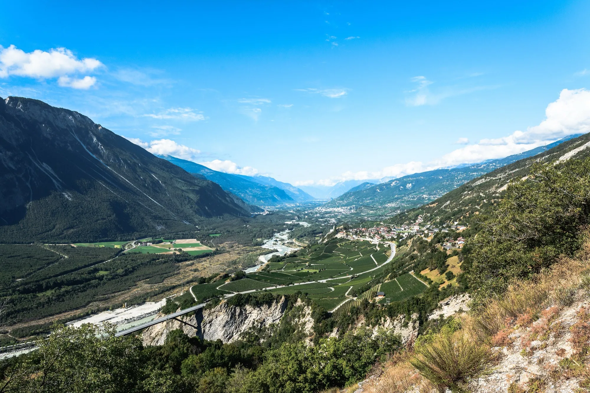Blick über Reben, ein Dorf und eine hohe Brücke hinunter ins breite Rhonetal
