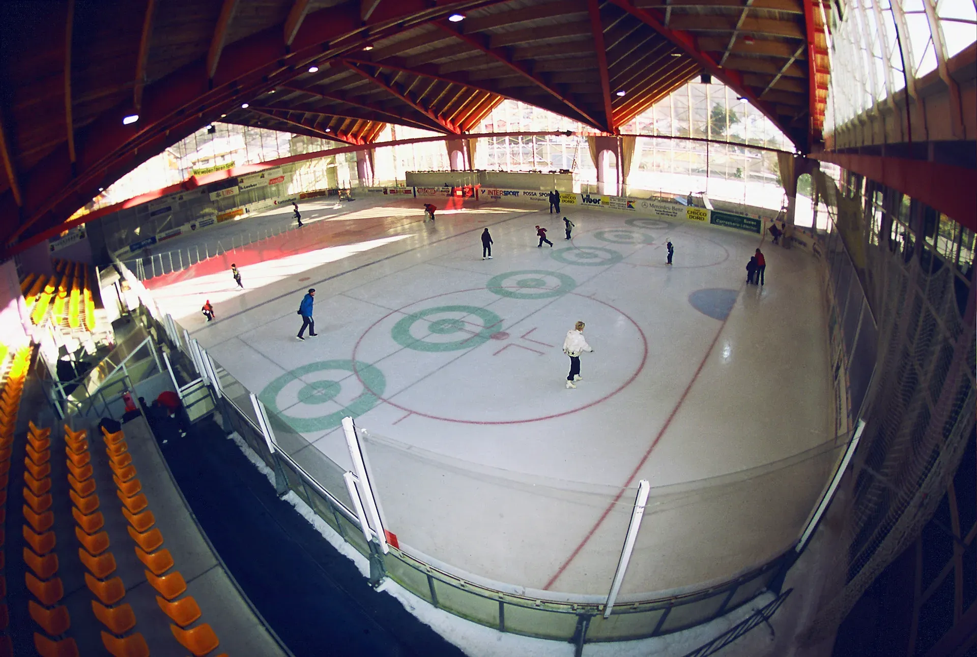 Hallen-Eisbahn mit wenigen Läufern, orangen Sitzen und einem roten Dach