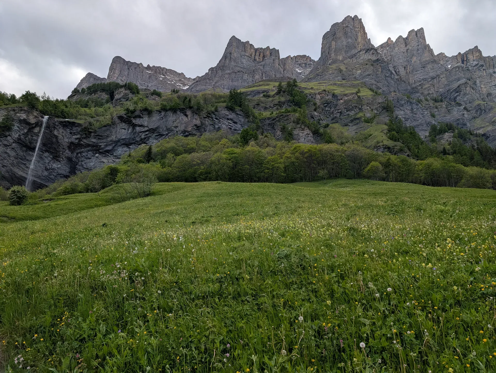 Blühende Wiese, links ein Wasserfall, dahinter gezackte Felswände