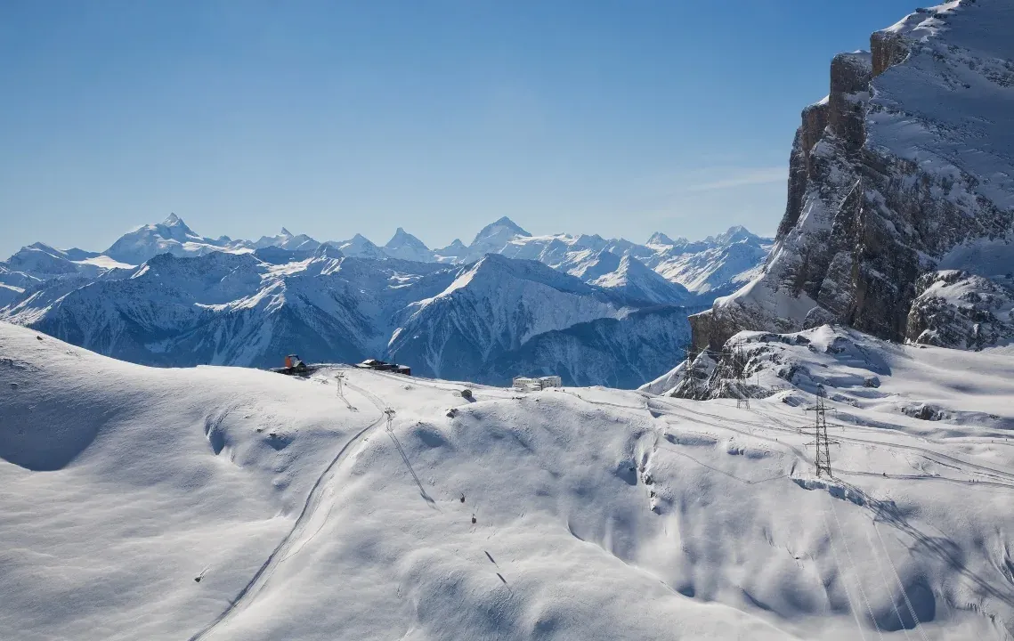 Verschneite Piste mit Liftspuren, rechts eine Felswand, im Hintergrund Gipfel