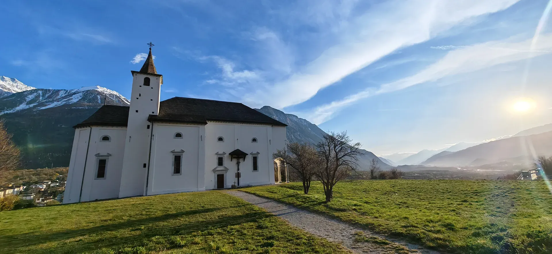 Weisse Kirche mit Glockenturm auf einer Wiese, Berge und Sonne dahinter