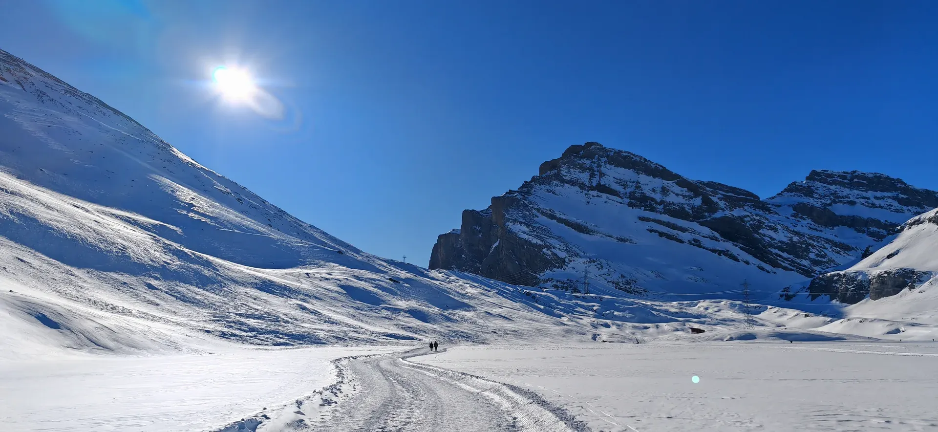 Zwei Personen auf einer Spur durch eine weite Schneefläche, rechts eine Felswand
