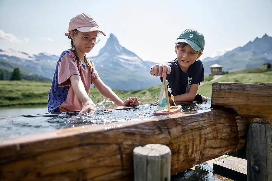 Zwei Kinder mit einem Spielschiff in einem Holztrog, dahinter das Matterhorn